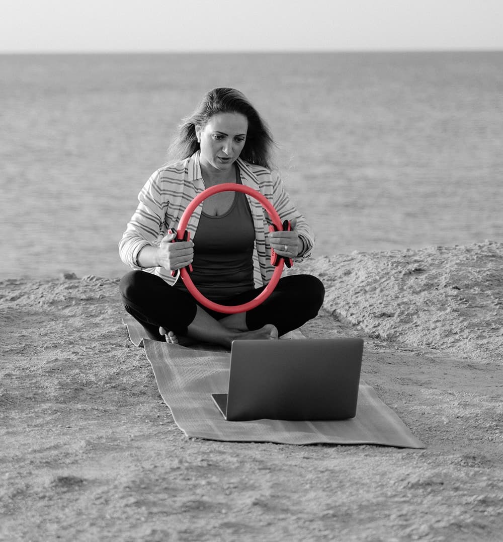 Nainen istuu jumppamatolla pilatesrengas käsissään ja seuraa treeniä kannettavalta tietokoneelta